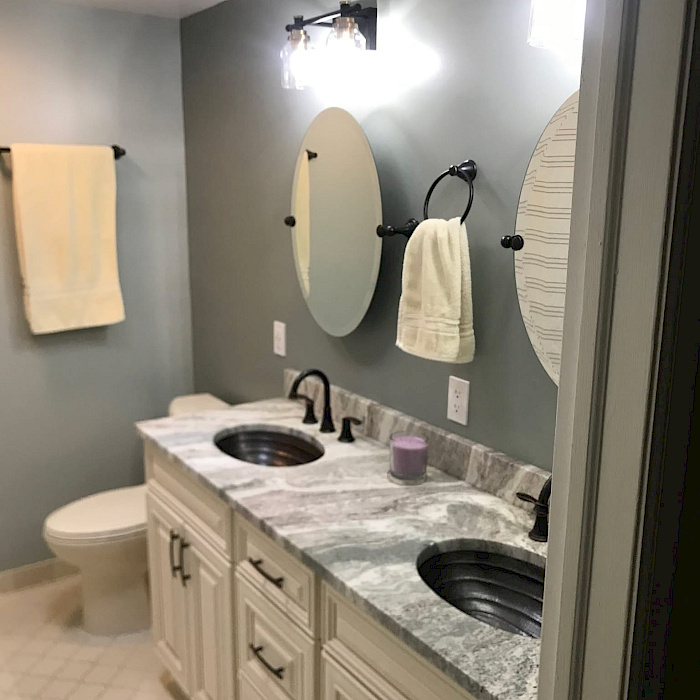 Double-vanity bathroom with hammered copper sinks, a marbled granite countertop, oval mirrors, and glass globe lighting.